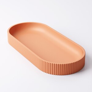 May include: A peach-colored, oval-shaped tray with a ribbed exterior. The tray has a smooth interior and is made of a solid material. The design is minimalist and modern, suitable for home decor or serving.