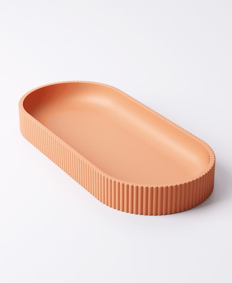 May include: A peach-colored, oval-shaped tray with a ribbed exterior. The tray has a smooth interior and is made of a solid material. The design is minimalist and modern, suitable for home decor or serving.