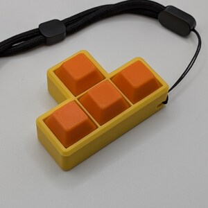 May include: A yellow and orange Tetris-shaped keychain with a black wrist strap. The keychain features six orange square buttons set in a yellow frame. The black strap has a black plastic clasp.