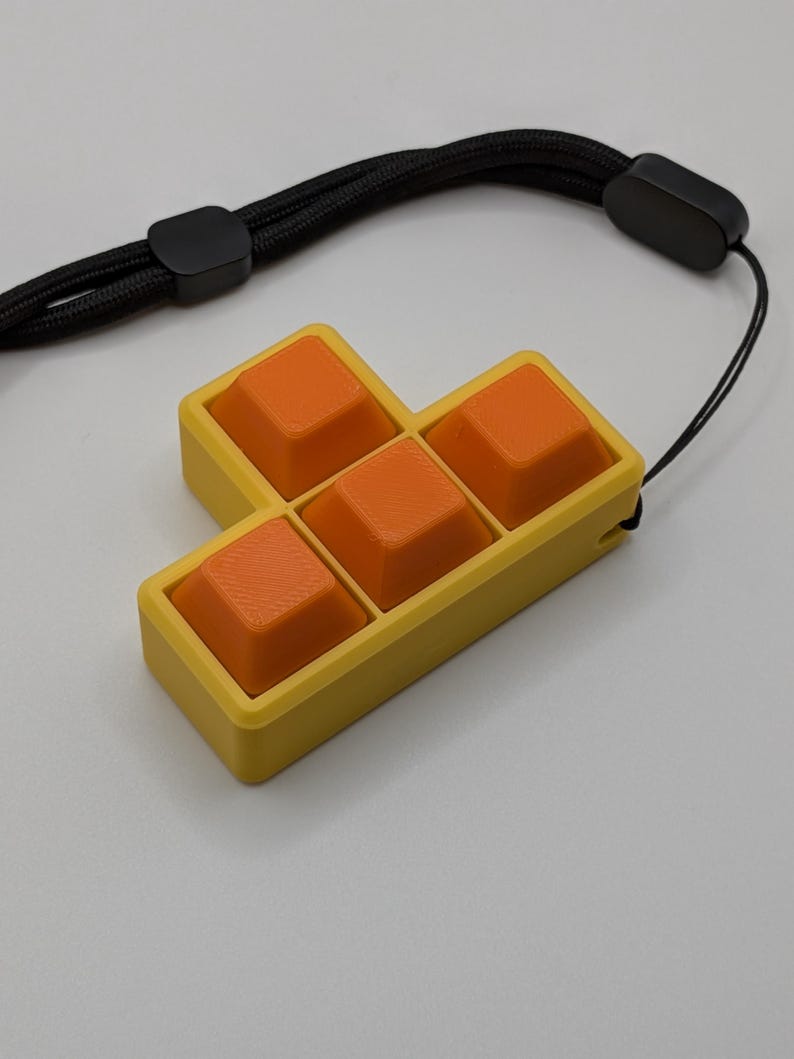 May include: A yellow and orange Tetris-shaped keychain with a black wrist strap. The keychain features six orange square buttons set in a yellow frame. The black strap has a black plastic clasp.