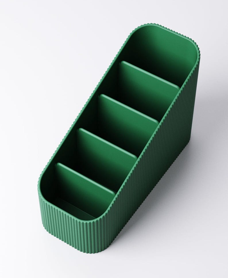 May include: A green desk organizer with five compartments. The organizer has a ribbed texture on the sides and a smooth interior. The compartments are separated by vertical dividers, and the overall shape is rectangular with rounded corners.
