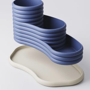 May include: A modern, abstract sculpture in shades of blue and off-white. The blue element features a tiered, wavy design with a cylindrical top and a curved, open section. It rests above a smooth, off-white, organically shaped tray.
