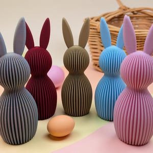 K&ouml;nnte beinhalten: Eine Sammlung farbenfroher, gerippter Hasenfiguren in Grau-, Burgunder-, Beige-, Blau- und Rosat&ouml;nen. Ostereier in Pastellfarben und ein Weidenkorb befinden sich im Hintergrund. Die Hasen haben lange Ohren und abgerundete K&ouml;rper.
