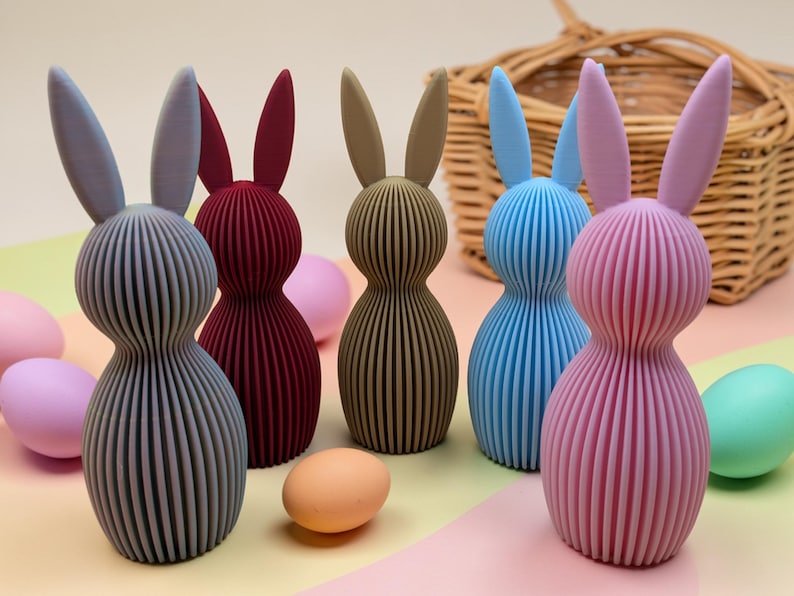 K&ouml;nnte beinhalten: Eine Sammlung farbenfroher, gerippter Hasenfiguren in Grau-, Burgunder-, Beige-, Blau- und Rosat&ouml;nen. Ostereier in Pastellfarben und ein Weidenkorb befinden sich im Hintergrund. Die Hasen haben lange Ohren und abgerundete K&ouml;rper.