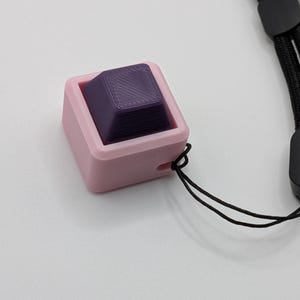 May include: A pink square-shaped object with a purple square inside, connected to a black lanyard. The purple square has a textured surface. The object is likely a fidget toy or a keycap.