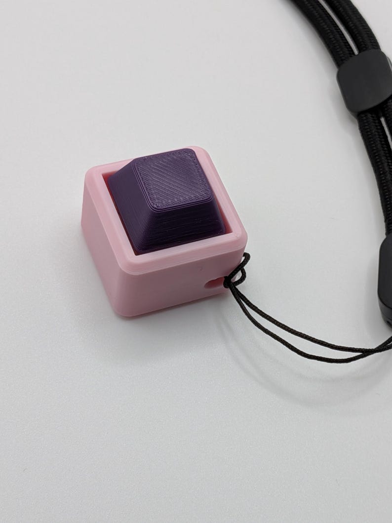 May include: A pink square-shaped object with a purple square inside, connected to a black lanyard. The purple square has a textured surface. The object is likely a fidget toy or a keycap.