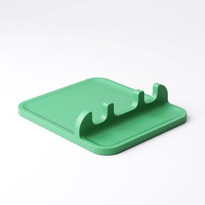 May include: A solid green spoon rest with a square base and three curved slots for utensils. The kitchen accessory is made of a matte material and is designed to keep work surfaces clean while cooking.