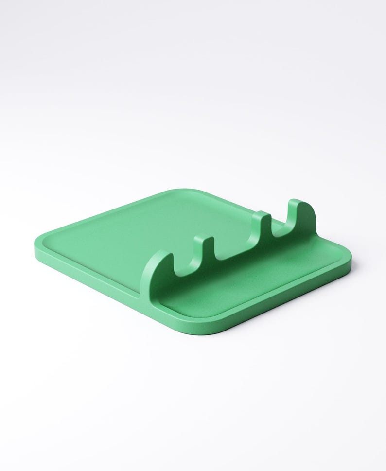 May include: A solid green spoon rest with a square base and three curved slots for utensils. The kitchen accessory is made of a matte material and is designed to keep work surfaces clean while cooking.