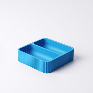 May include: A bright blue, rectangular container with two compartments. The container has a ribbed exterior and is set against a white background. The design is modern and minimalist, suitable for organizing small items.