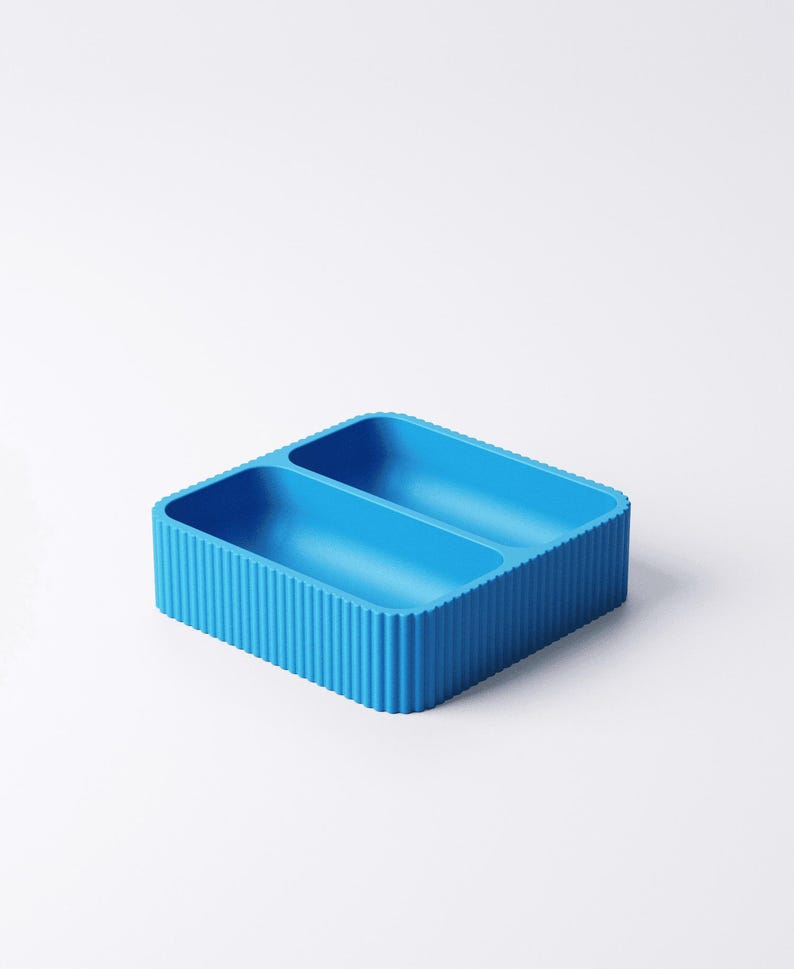 May include: A bright blue, rectangular container with two compartments. The container has a ribbed exterior and is set against a white background. The design is modern and minimalist, suitable for organizing small items.
