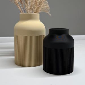 May include: Two decorative vases, one beige and one black, sit side-by-side. The beige vase is tall and cylindrical, with vertical ribbed detailing on the lower half. The black vase is shorter and has a similar ribbed design. Dried ornamental grass is in the beige vase.