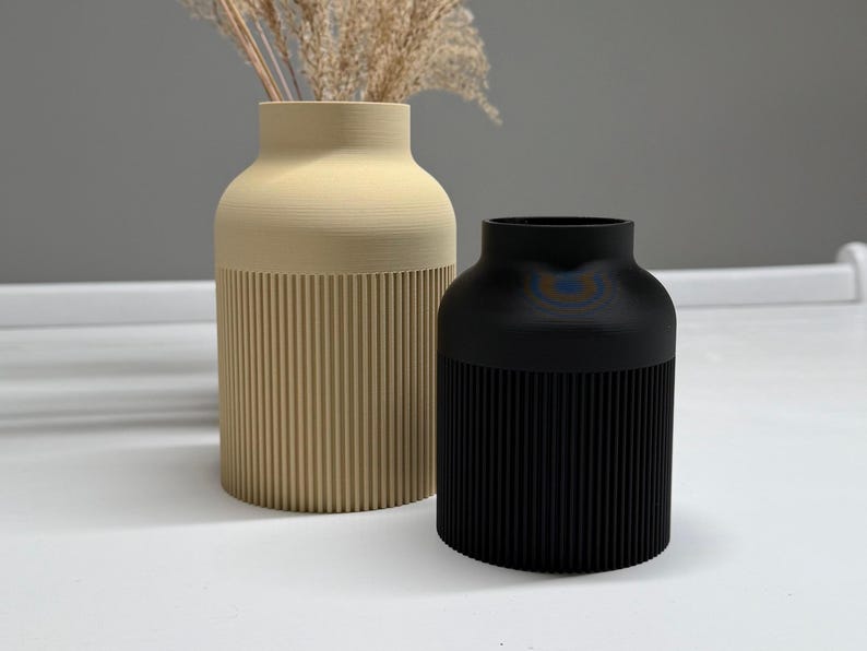 May include: Two decorative vases, one beige and one black, sit side-by-side. The beige vase is tall and cylindrical, with vertical ribbed detailing on the lower half. The black vase is shorter and has a similar ribbed design. Dried ornamental grass is in the beige vase.
