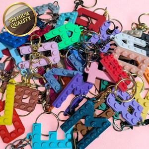 May include: A collection of colorful letter-shaped keychains, resembling building blocks, with gold-colored key rings and clasps. The keychains are in various colors, including red, green, blue, and purple, against a pink background. A gold-rimmed sticker reads "Premium Quality."