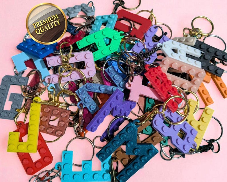 May include: A collection of colorful letter-shaped keychains, resembling building blocks, with gold-colored key rings and clasps. The keychains are in various colors, including red, green, blue, and purple, against a pink background. A gold-rimmed sticker reads "Premium Quality."