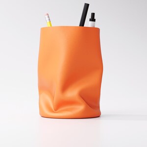 May include: An orange desk organizer with a unique, crumpled design. The container holds a yellow pencil, a black pen, and a white marker. The organizer is made of a soft material and sits on a white surface.