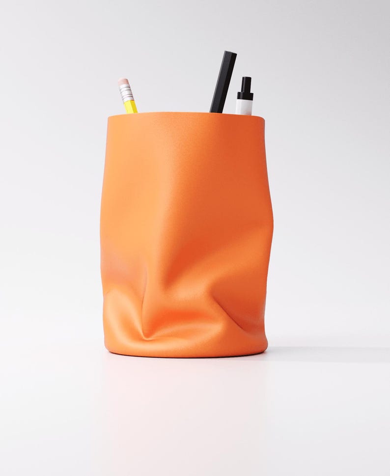 May include: An orange desk organizer with a unique, crumpled design. The container holds a yellow pencil, a black pen, and a white marker. The organizer is made of a soft material and sits on a white surface.