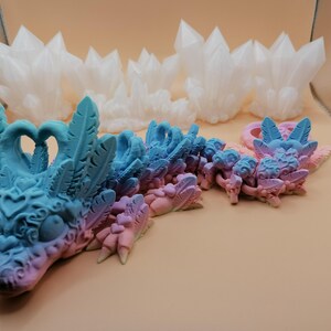 May include: A detailed image of a fantasy dragon sculpture in shades of blue and pink, with intricate feather and heart details. The dragon is positioned in front of several white crystal formations, creating a whimsical and artistic display.