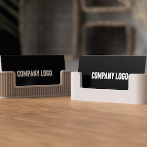 May include: Two business card holders on a wooden surface. Each holder is a different design, one with vertical ridges and the other with a smooth, rounded shape. Both hold black cards with the text "COMPANY LOGO" in white.