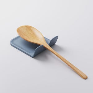 Op de afbeelding: Een houten lepel rust op een blauwe lepelhouder. De lepel is lichtbruin met een lange steel en een afgeronde kom. De lepelhouder is effen blauw en heeft een gebogen ontwerp om de lepel vast te houden.