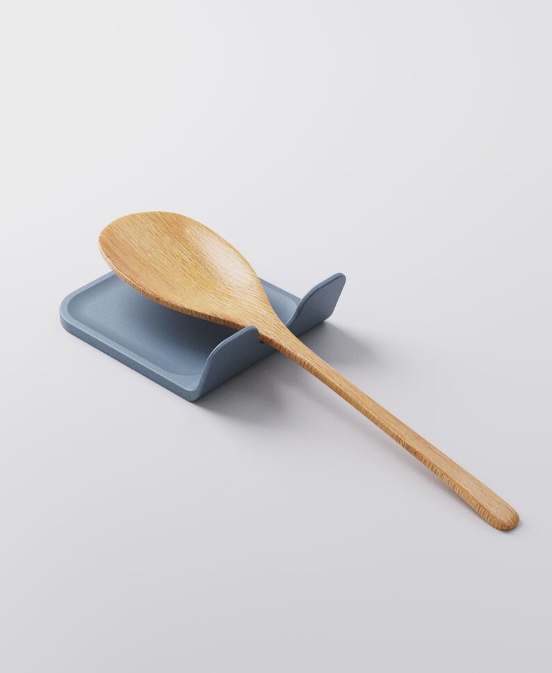 Op de afbeelding: Een houten lepel rust op een blauwe lepelhouder. De lepel is lichtbruin met een lange steel en een afgeronde kom. De lepelhouder is effen blauw en heeft een gebogen ontwerp om de lepel vast te houden.