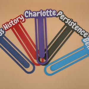 May include: Five large, flat, paperclip-shaped bookmarks in blue, red, purple, dark grey, and light blue. Each bookmark features a word or name in white lettering with a dark outline: Focus, History, Charlotte, Persistence, and Believe.