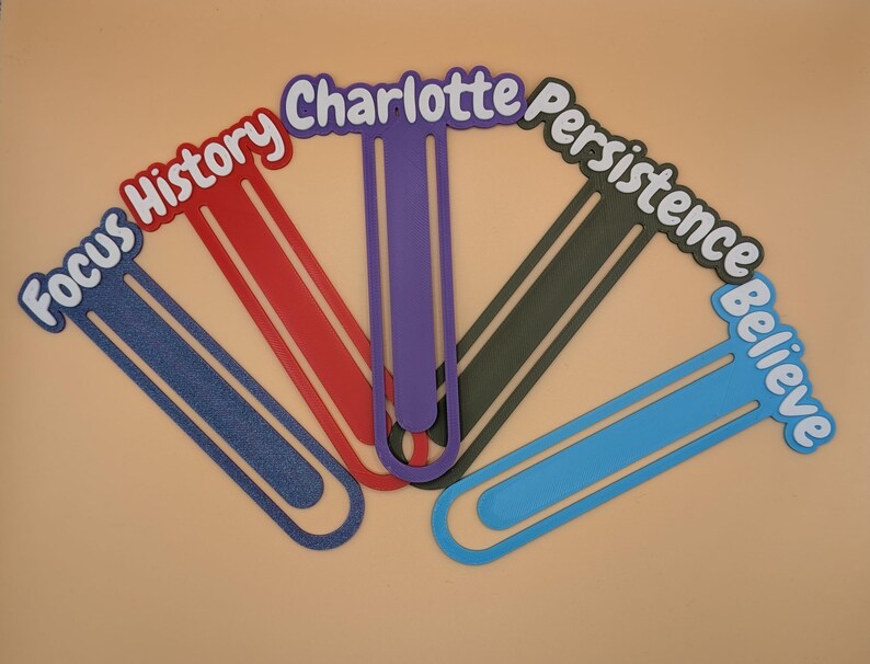 May include: Five large, flat, paperclip-shaped bookmarks in blue, red, purple, dark grey, and light blue. Each bookmark features a word or name in white lettering with a dark outline: Focus, History, Charlotte, Persistence, and Believe.
