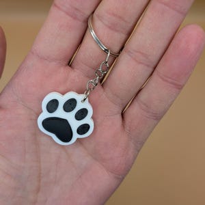 May include: A white and black paw print keychain. The keychain features a paw print design with a white background and black paw pads. The keychain is attached to a silver keyring.