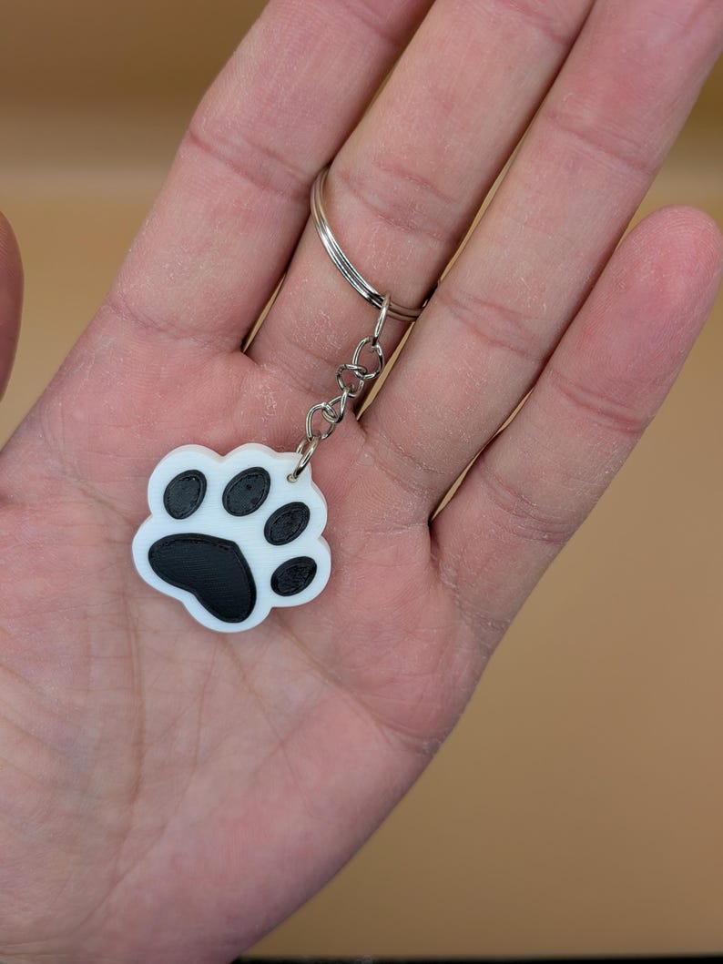 May include: A white and black paw print keychain. The keychain features a paw print design with a white background and black paw pads. The keychain is attached to a silver keyring.