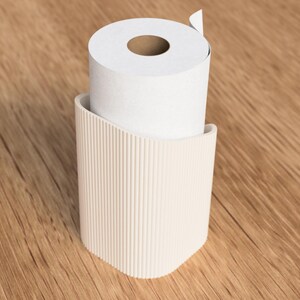 Op de afbeelding: Een witte rol toiletpapier staat in een beige, geribbelde houder. De houder heeft een gebogen ontwerp en is gemaakt van een solide materiaal. De achtergrond is een lichtbruin houten oppervlak.