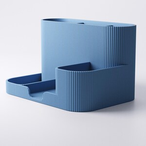 May include: A blue desk tidy with a ribbed texture. It features a tall, rectangular section, a smaller, curved section, and a tray with two compartments. The design is modern and minimalist.