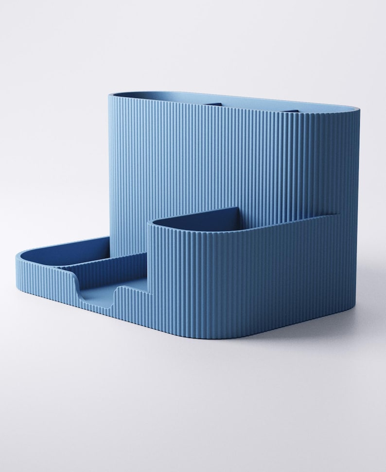 May include: A blue desk tidy with a ribbed texture. It features a tall, rectangular section, a smaller, curved section, and a tray with two compartments. The design is modern and minimalist.