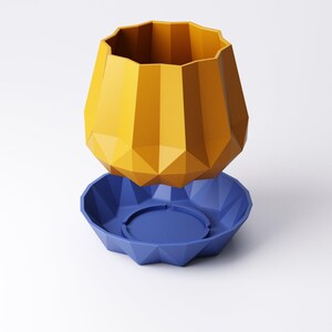 May include: A geometric, two-piece planter set. The top piece is a faceted, mustard-yellow vase. The base is a faceted, deep blue saucer with a circular indentation in the center. The planter is designed with a modern aesthetic.
