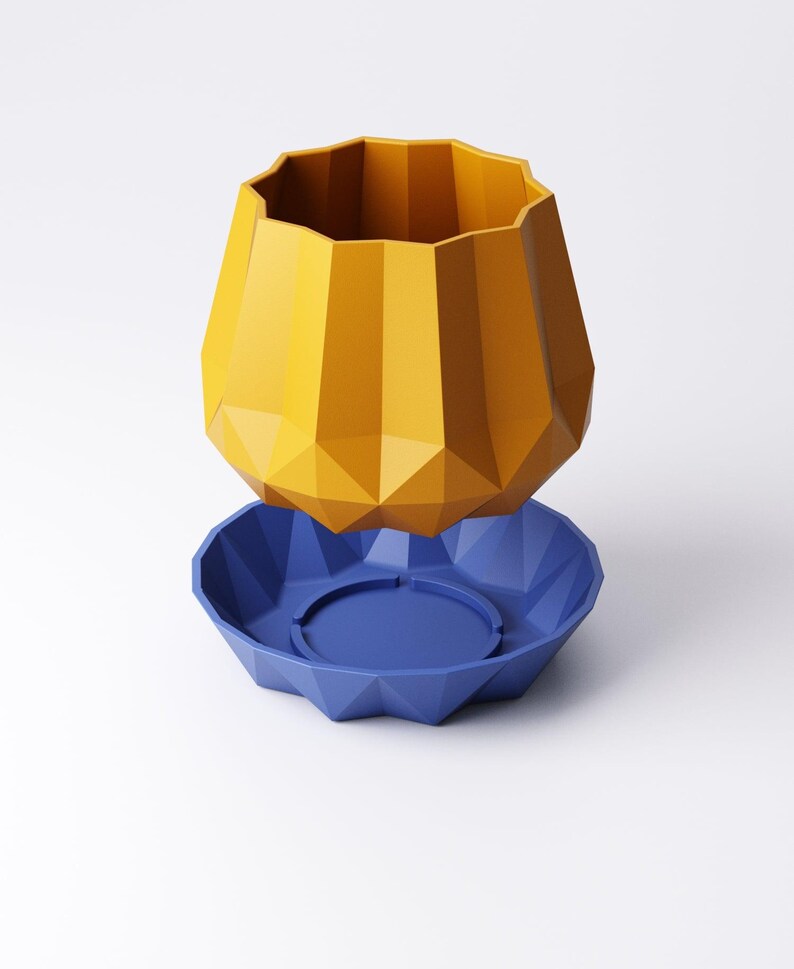 May include: A geometric, two-piece planter set. The top piece is a faceted, mustard-yellow vase. The base is a faceted, deep blue saucer with a circular indentation in the center. The planter is designed with a modern aesthetic.