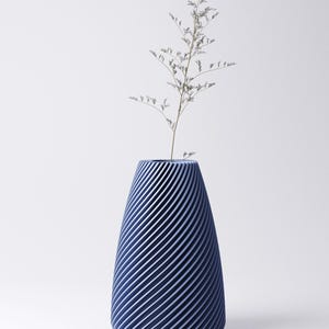 K&ouml;nnte beinhalten: Eine marineblaue Vase mit Spiralmuster, die eine schlanke, getrocknete Pflanze enth&auml;lt. Die Vase hat eine konische Form mit diagonalen Linien, die ein strukturiertes Aussehen erzeugen. Der Hintergrund ist schlicht wei&szlig;.