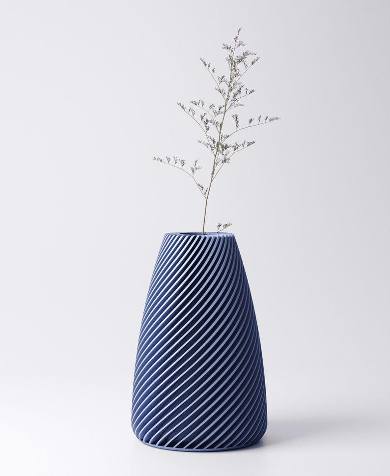 K&ouml;nnte beinhalten: Eine marineblaue Vase mit Spiralmuster, die eine schlanke, getrocknete Pflanze enth&auml;lt. Die Vase hat eine konische Form mit diagonalen Linien, die ein strukturiertes Aussehen erzeugen. Der Hintergrund ist schlicht wei&szlig;.