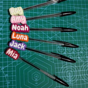 May include: Six clear ballpoint pens with black ink, each topped with a colorful name tag. The names on the tags are Roy, Amy, Noah, Luna, Jack, and Mia. The pens are arranged on a green cutting mat.