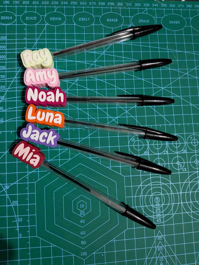 May include: Six clear ballpoint pens with black ink, each topped with a colorful name tag. The names on the tags are Roy, Amy, Noah, Luna, Jack, and Mia. The pens are arranged on a green cutting mat.