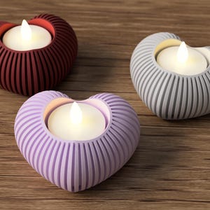 May include: Three heart-shaped candle holders in red, gray, and lavender. Each holder has a ribbed design and holds a small, glowing, white candle. The holders are arranged on a wooden surface, creating a warm and inviting ambiance.
