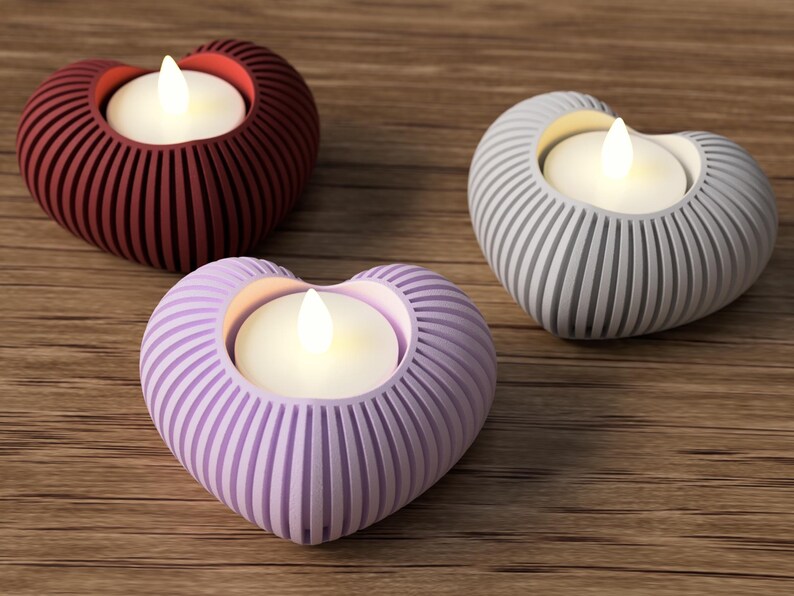 May include: Three heart-shaped candle holders in red, gray, and lavender. Each holder has a ribbed design and holds a small, glowing, white candle. The holders are arranged on a wooden surface, creating a warm and inviting ambiance.