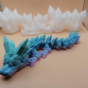 May include: A 3D-printed articulated dragon in shades of blue and purple, with crystal-like spikes along its back and head. Several white crystal formations are in the background. The dragon is on a light brown surface.