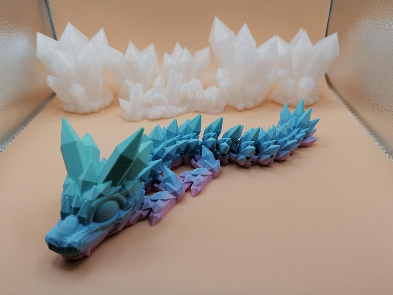 May include: A 3D-printed articulated dragon in shades of blue and purple, with crystal-like spikes along its back and head. Several white crystal formations are in the background. The dragon is on a light brown surface.