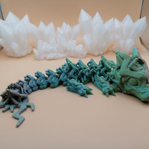 May include: A 3D-printed articulated dragon toy in shades of green and blue, with a detailed head and segmented body. Several white, crystalline formations are in the background, suggesting a fantasy theme.
