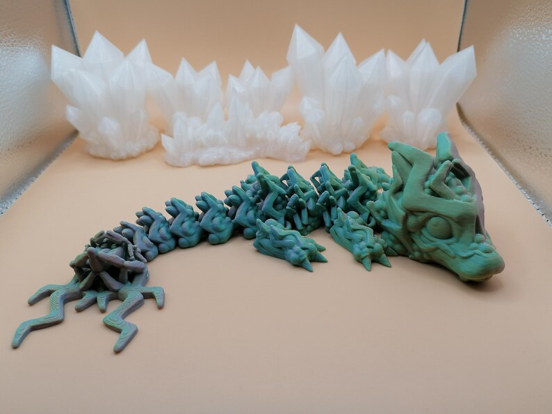 May include: A 3D-printed articulated dragon toy in shades of green and blue, with a detailed head and segmented body. Several white, crystalline formations are in the background, suggesting a fantasy theme.
