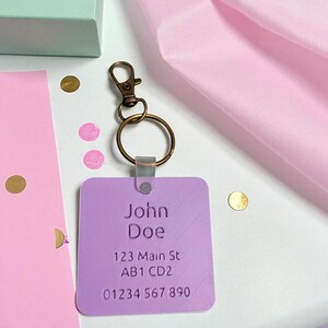 May include: A lavender rectangular keychain with the name "John Doe", address, and phone number printed in white. The keychain has a bronze-colored clasp and ring. The background includes confetti and pink tissue paper.