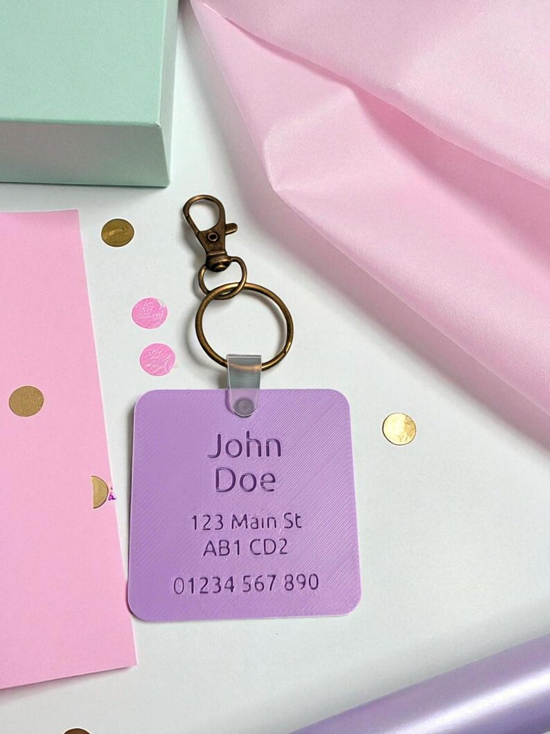 May include: A lavender rectangular keychain with the name "John Doe", address, and phone number printed in white. The keychain has a bronze-colored clasp and ring. The background includes confetti and pink tissue paper.