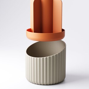 May include: A two-piece decorative container set. The top piece is a terracotta-colored, open-top cylinder with vertical dividers. The bottom piece is a light gray, ribbed, semi-circular container. The pieces are designed to fit together.