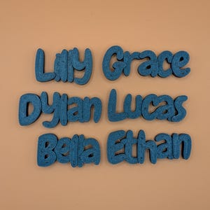 May include: Six teal wooden name cutouts: Lilly, Grace, Dylan, Lucas, Bella, and Ethan. The names are in a playful, rounded font, and are arranged in two rows on a peach-coloured background.