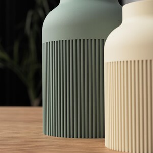 May include: Two cylindrical vases, one sage green and the other cream, stand side-by-side on a wooden surface. The lower portion of each vase features vertical, ridged detailing. The background is blurred, with hints of greenery and a dark wall.