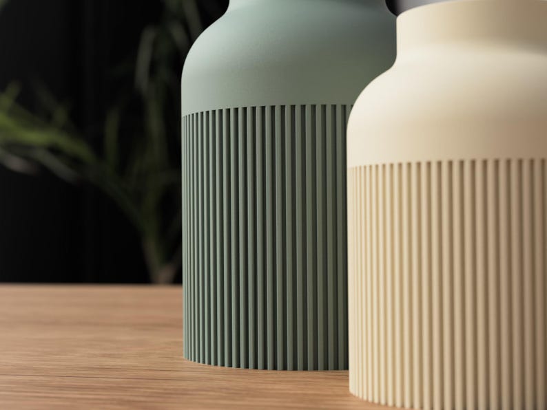 May include: Two cylindrical vases, one sage green and the other cream, stand side-by-side on a wooden surface. The lower portion of each vase features vertical, ridged detailing. The background is blurred, with hints of greenery and a dark wall.