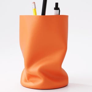 May include: An orange desk organizer with a unique, crumpled design. The container holds a yellow pencil, a black pencil, and a black and white pen. The organizer is made of a smooth material and sits on a white surface.